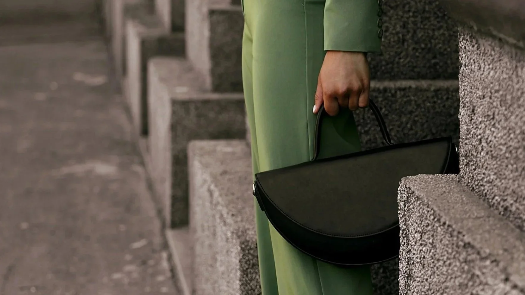 Une personne portant un costume vert et tenant un sac à main noir se tient près d'un mur en pierre.