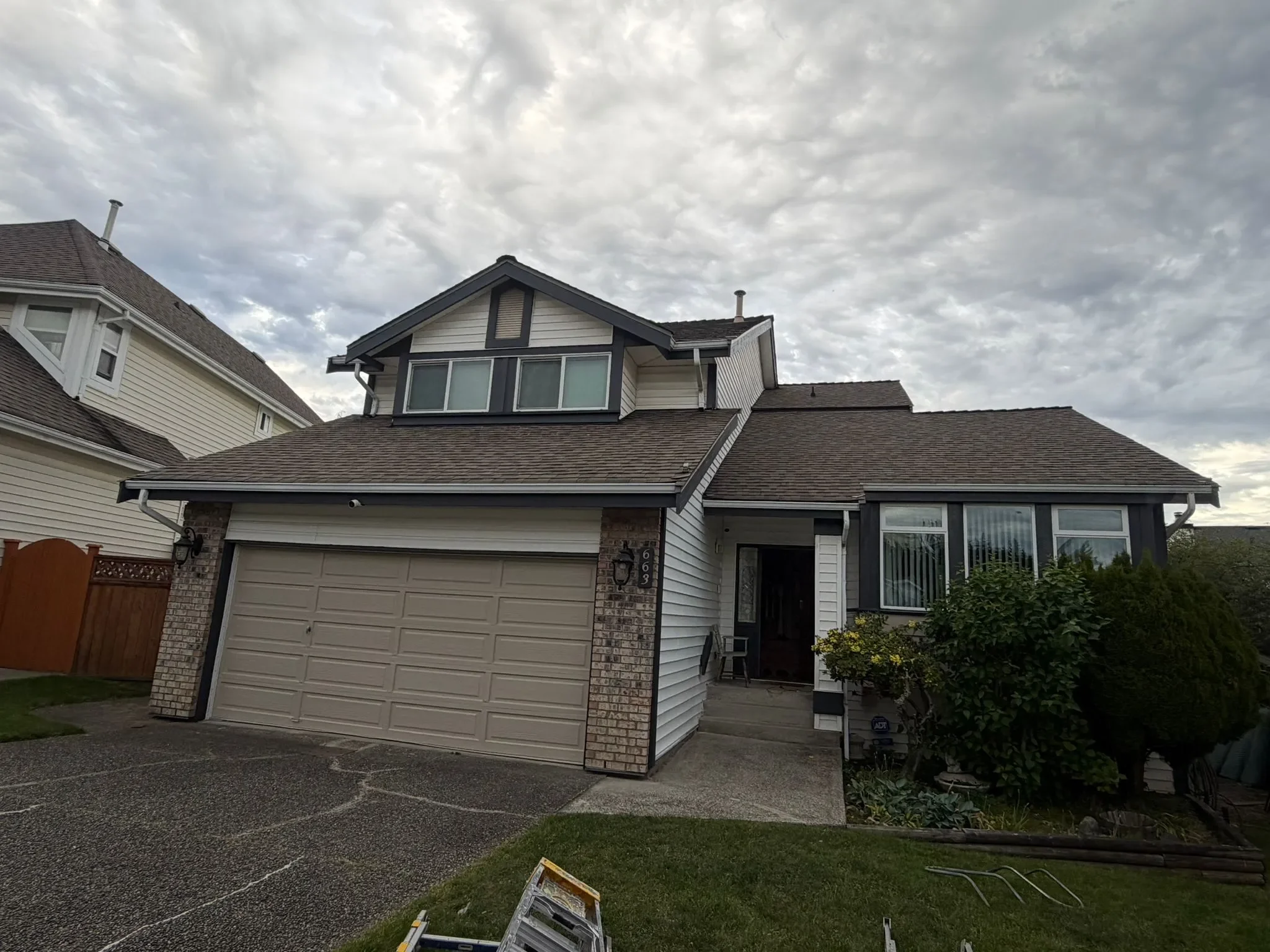 A two-story suburban house with a garage door, brick accents, and a front porch in West Vancouver. The sky above is cloudy.