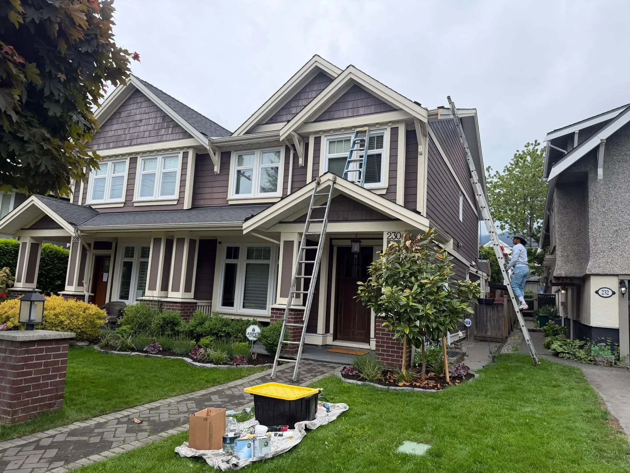 A two-story house with purple siding in Vancouver, white trim, and multiple windows is undergoing painting, with a ladder leaning against the upper floor. A worker is on a ladder on the right side of the house, working on the exterior. There are pain