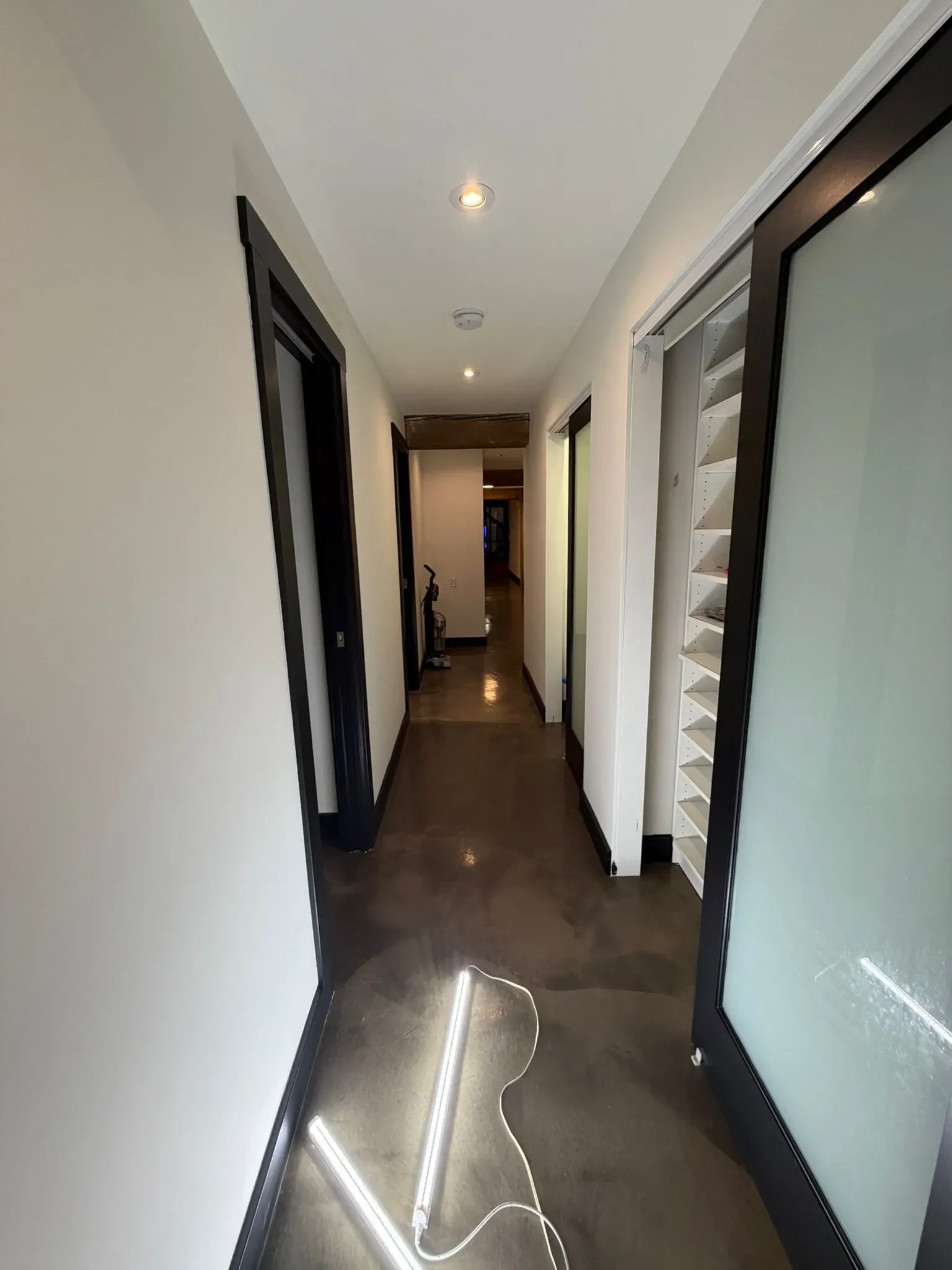 A hallway with black door frames, white walls, and a polished dark floor, with LED lighting installed on the ceiling and a tube light on the floor in Burnaby.