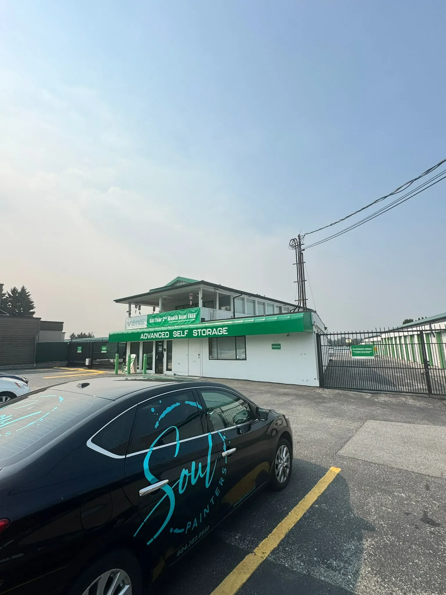 Parking lot in front of a two-story self-storage facility with a green sign that reads 'Advanced Self Storage'. A black car with blue and white branding for 'Soul Painters' is parked in the foreground, with a cloudy sky overhead.