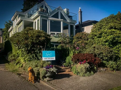 A large, multi-story house with multiple balconies and a glass-enclosed upper floor, surrounded by lush bushes and flowering plants. A sign with the word 'Salon' and a phone number is in the front yard, and a small dog is sitting on the sidewalk nearby.