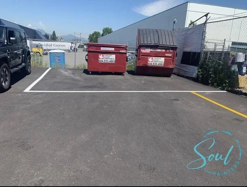 Two red dumpsters in an empty parking lot next to chain-link fence, building and yellow curb in background, maintained by painters offering exterior painting services and free estimate near you.