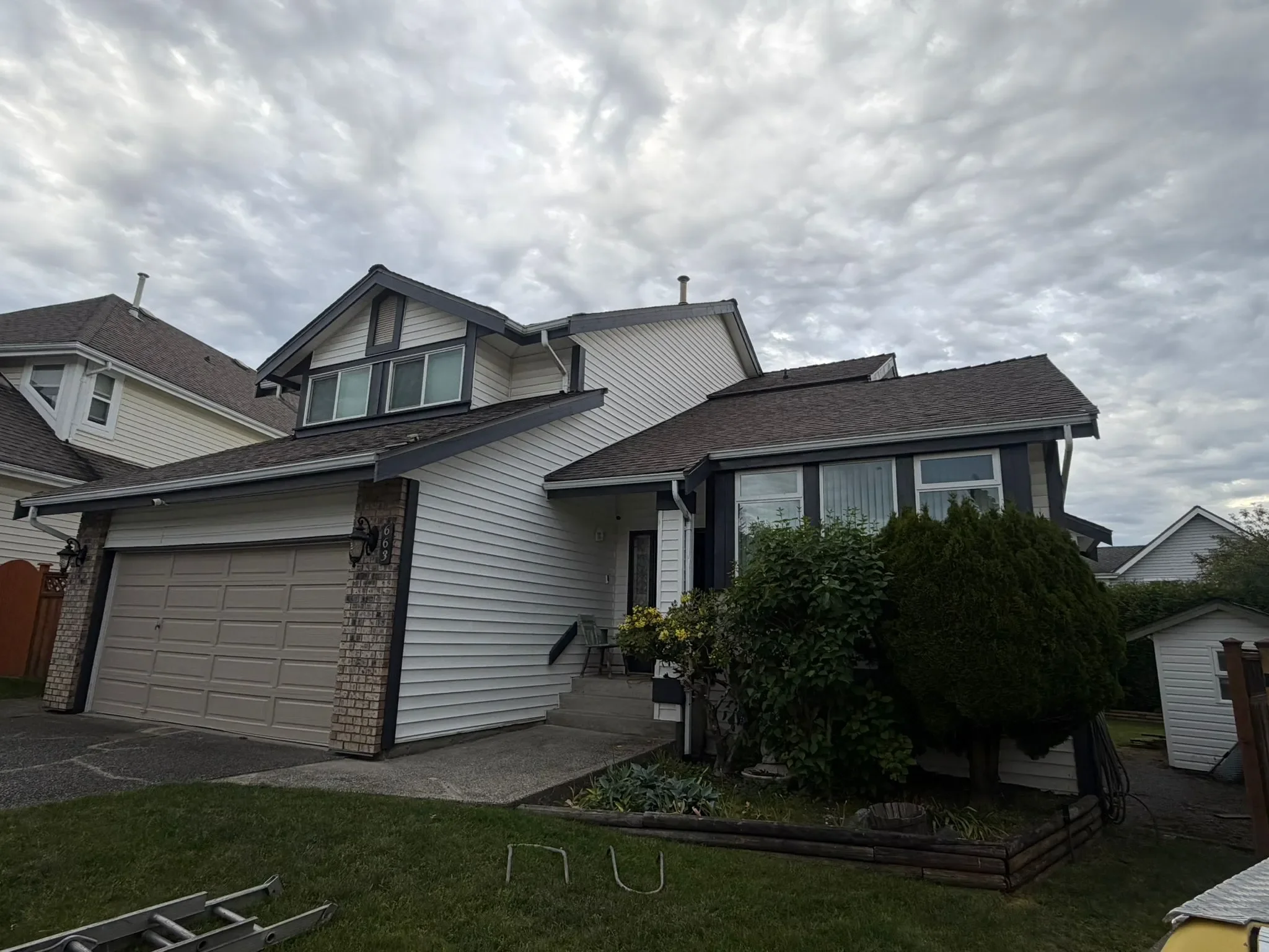 A two-story residential house with a white exterior and a gray roof, featuring multiple windows and a garage door in West Vancouver. The front yard has a small lawn, garden, and outdoor furniture. The sky is cloudy.
