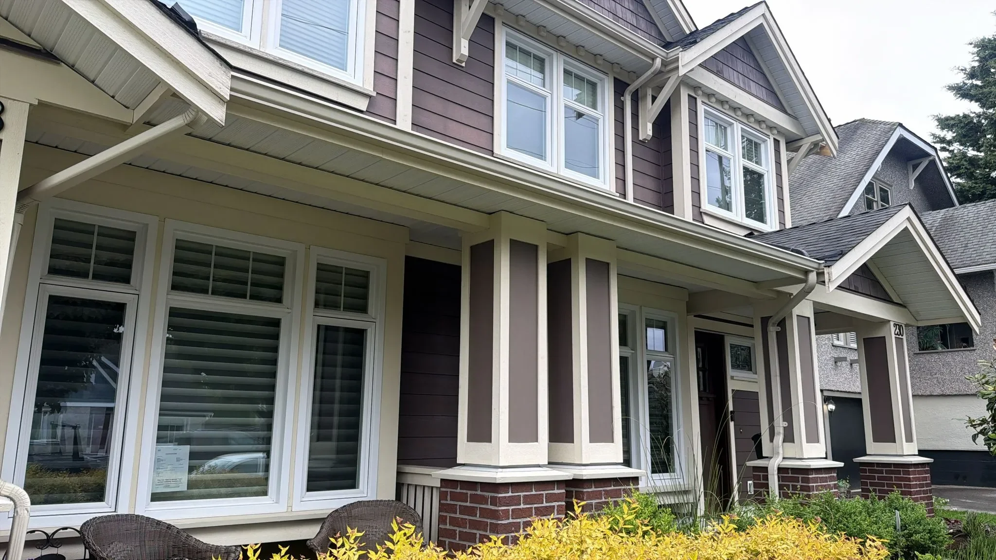 A two-story suburban house with gray and white siding in Vancouver, multiple large windows, a covered front porch, and a brick foundation. There are yellow bushes in the front yard and a patio area with chairs.
