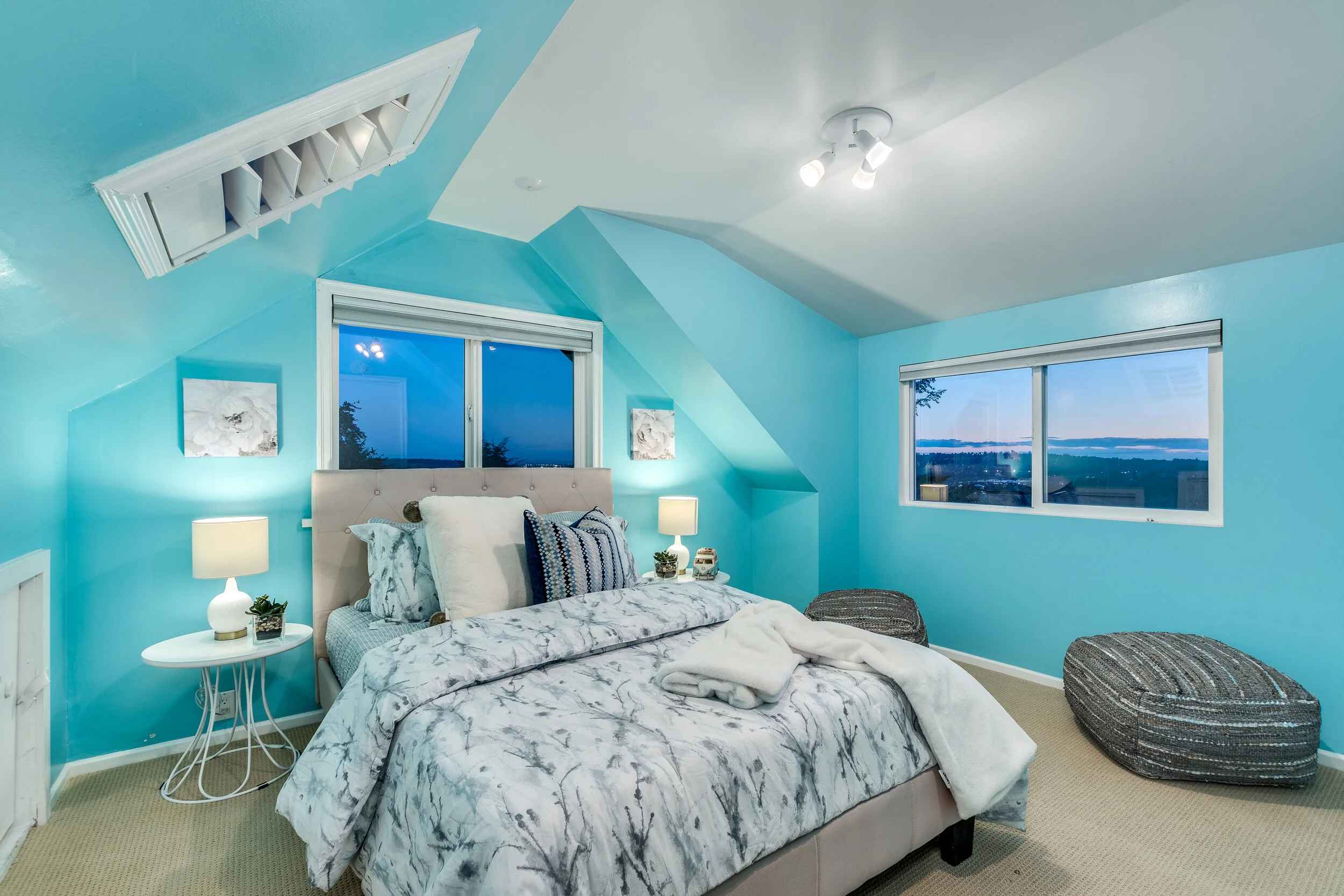 A cozy bedroom with light blue walls, a bed with marble-patterned bedding, decorative pillows, and a white blanket. It has two windows showing an evening view of the sky and landscape. There are two bedside lamps, wall art, an air vent, and two textured ottomans on a beige carpet.