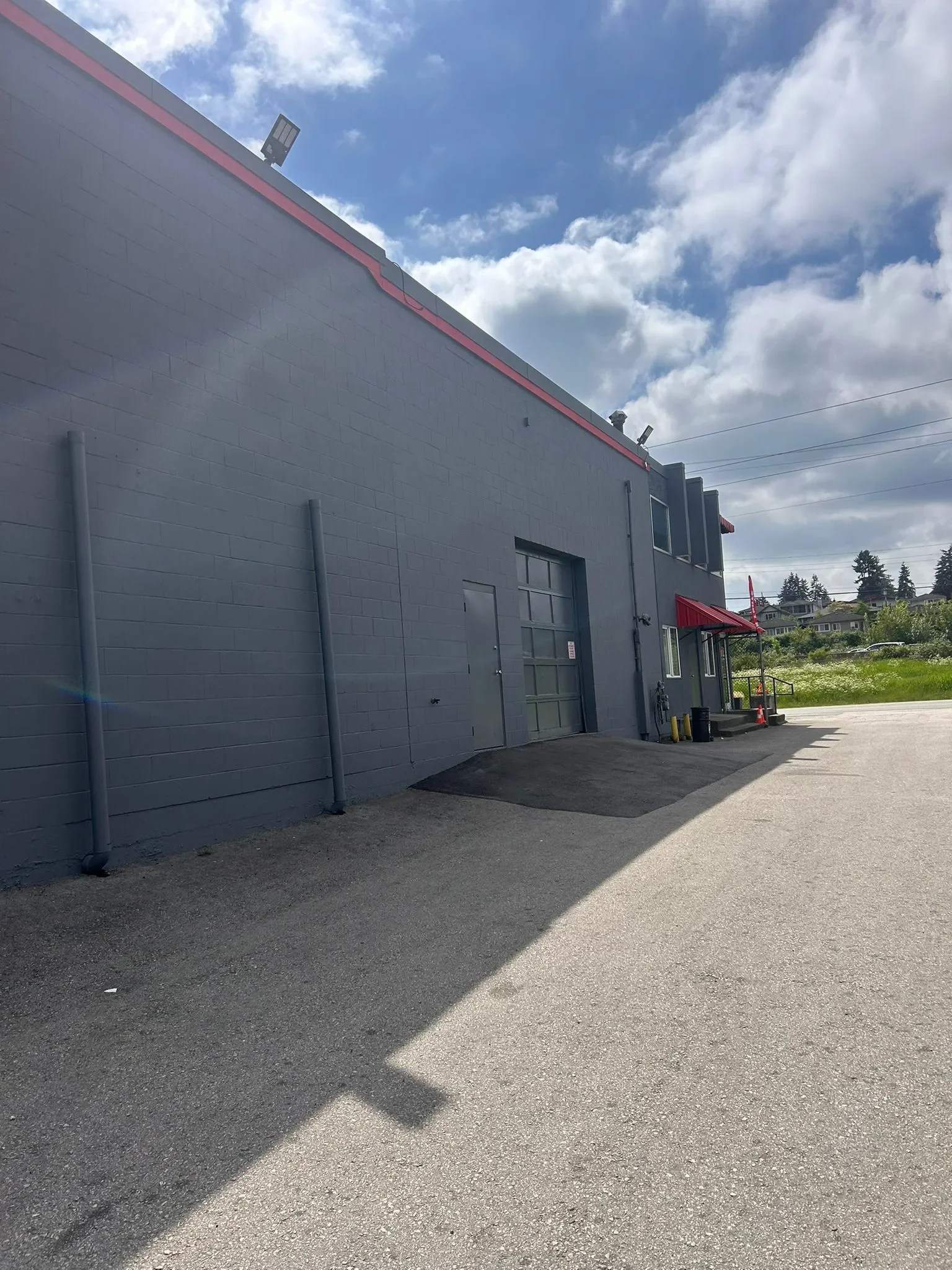A gray commercial building with a red trim on the roof, a gray garage door, and a sidewalk with an orange cone and some yellow bollards. The sky is partly cloudy with blue sky and white clouds.