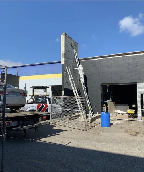 Painter spray painting a wall black on a ladder outside a commercial building, with parked cars and construction supplies, offering free painting estimate and exterior painting services near you.