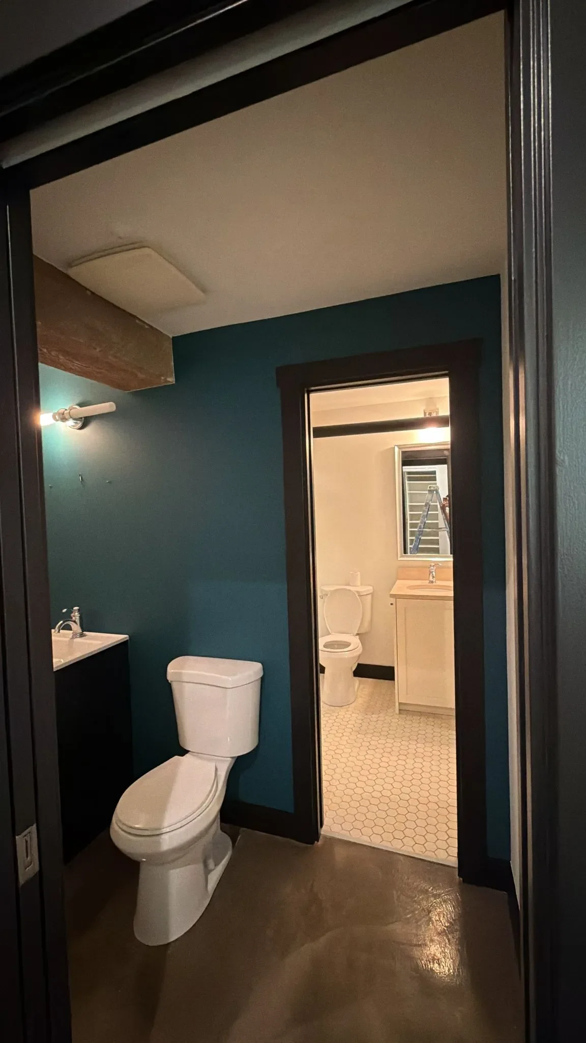 View of a bathroom with a dark green wall, a white toilet, a vanity with a sink, and an adjacent room featuring a mirror, a toilet, and a small cabinet in New Westminster.