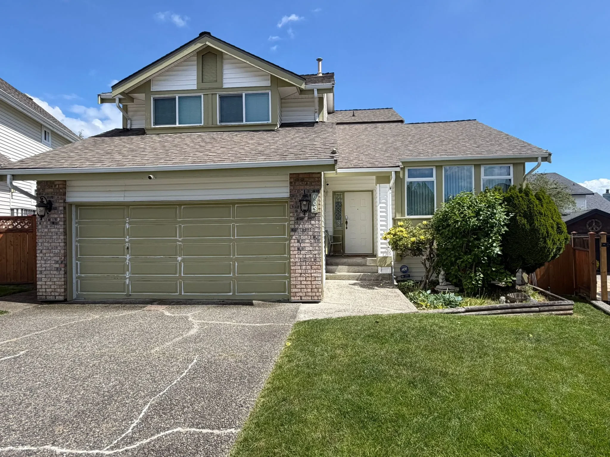 A two-story house with a grey garage door, white siding, brick accents, and a front door with a small window in a suburban neighborhood in West Vancouver. There is a garage, steps leading to the front door, a small tree, and well-maintained yard.