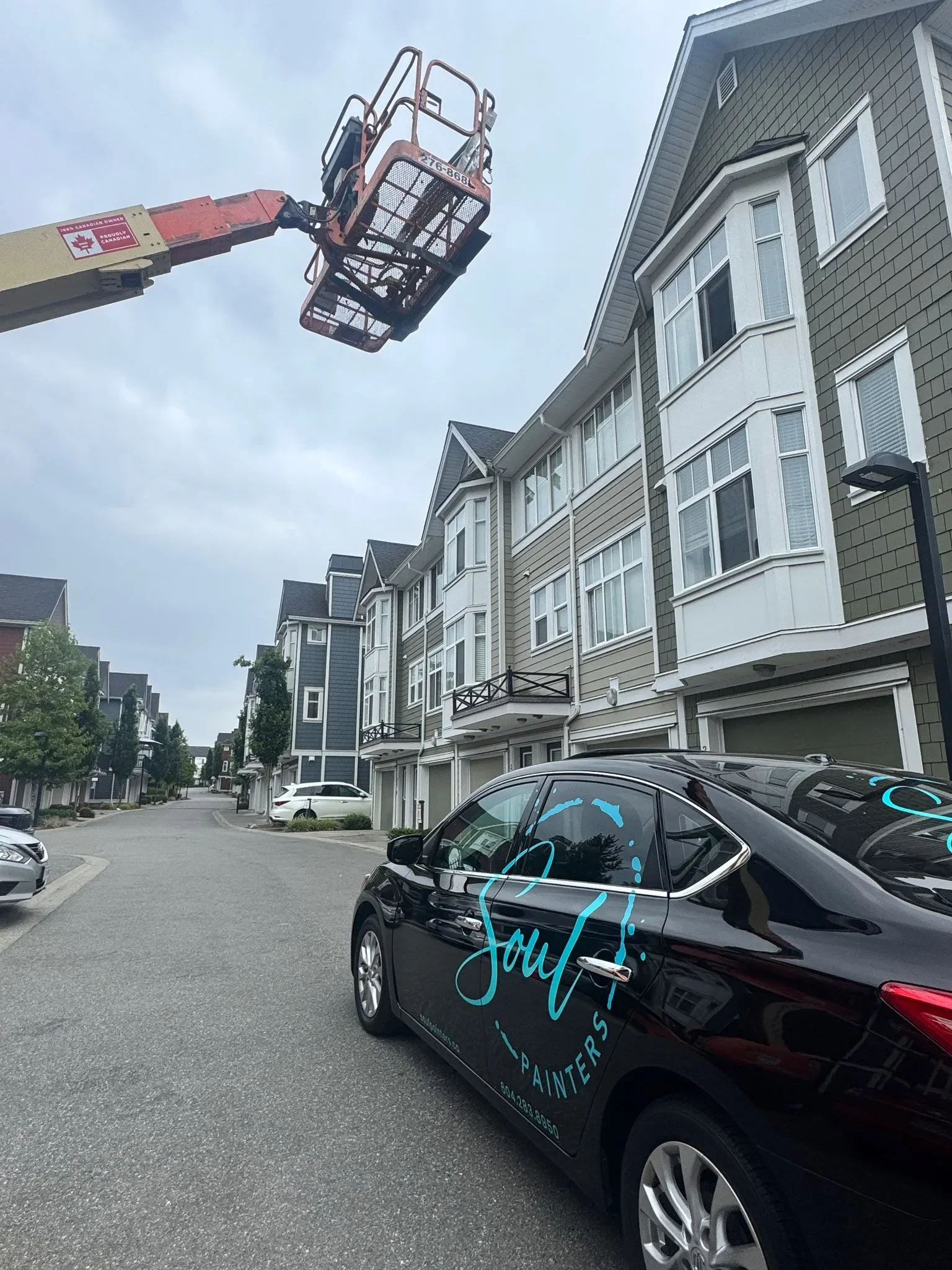 A black car parked on a residential street with apartment buildings. A cherry picker lift is extended overhead. The car has a blue and white logo for a painting company called 'Soul Painters'.