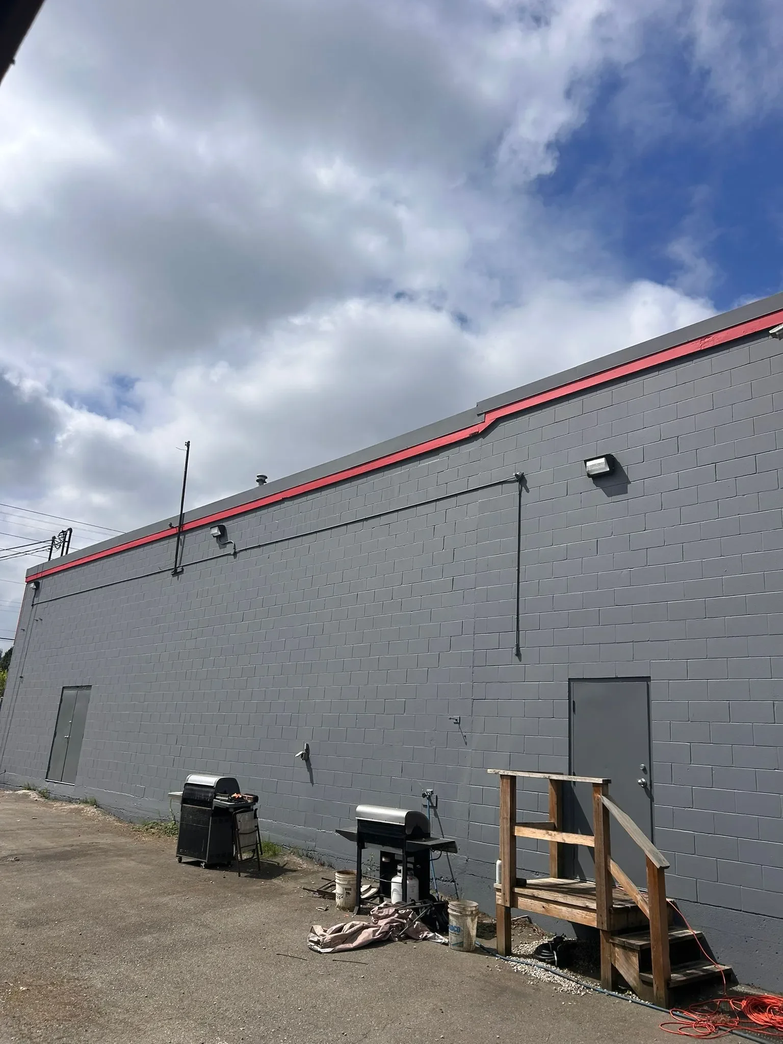 The side of a gray building with construction tools and materials around it, including a black trash bin, a small wooden staircase, buckets, and electrical cords.