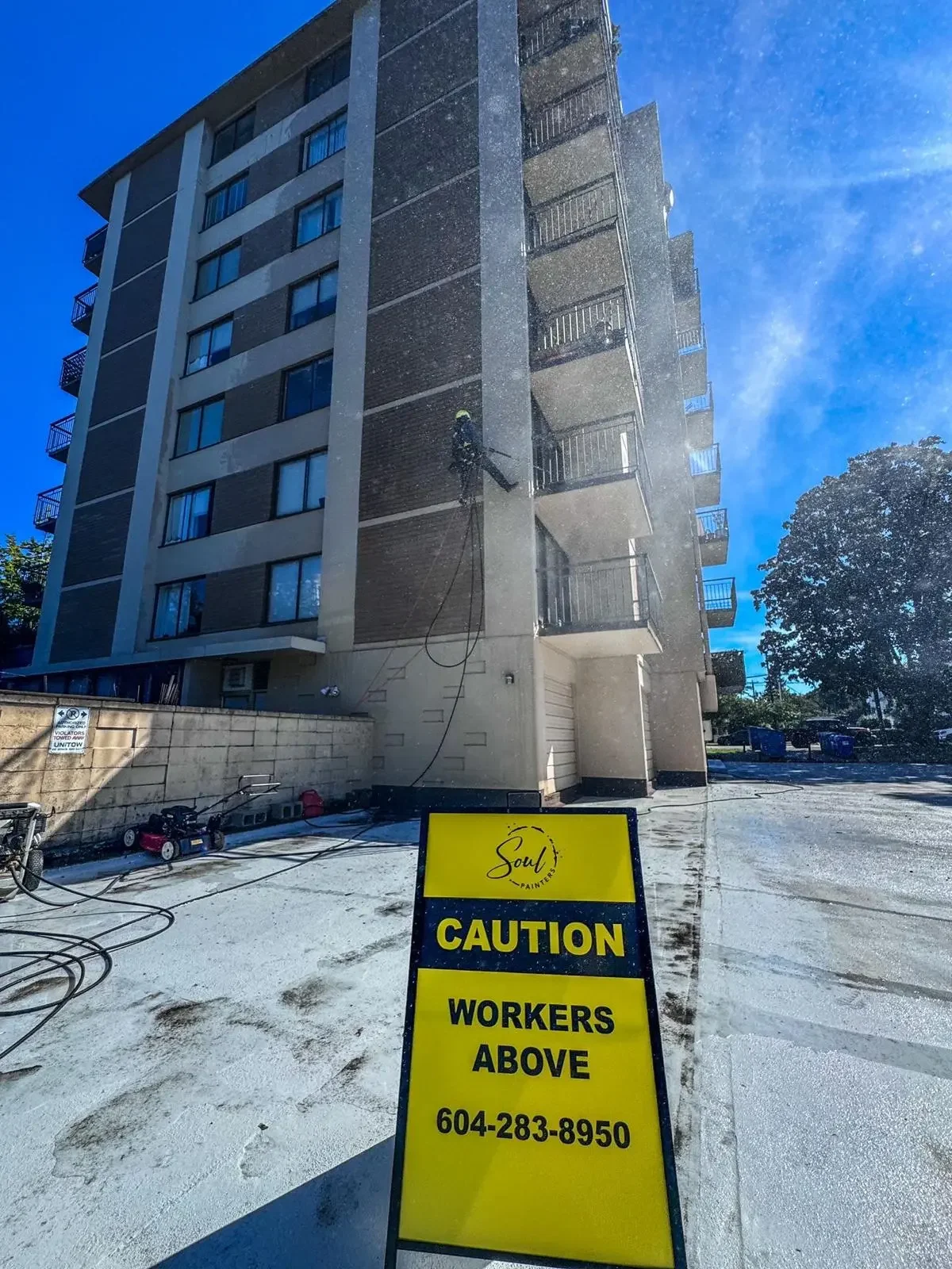 A worker cleaning the exterior of a high-rise apartment building with water spray. A yellow caution sign in the foreground warns of workers above, with contact info for Soul Painters.