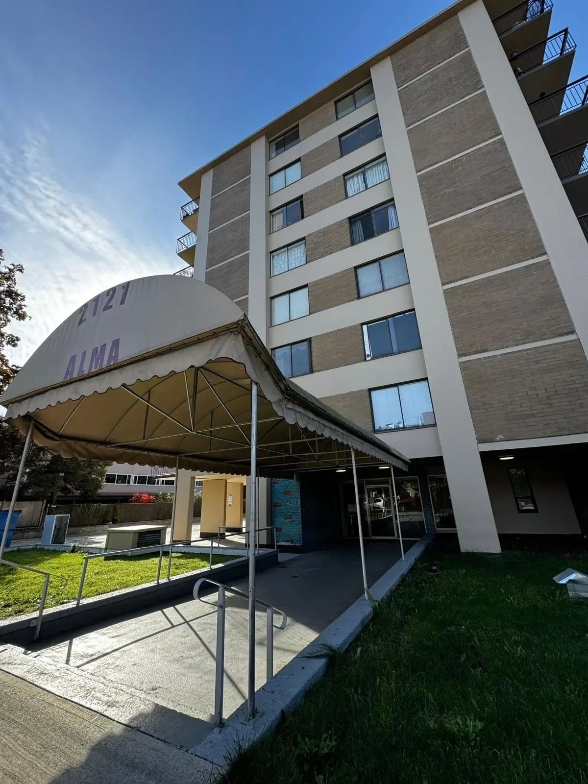 Exterior view of a multi-story apartment building with a small lawn and a covered entrance area labeled '2121 ALMA', taken during daytime with clear blue sky.