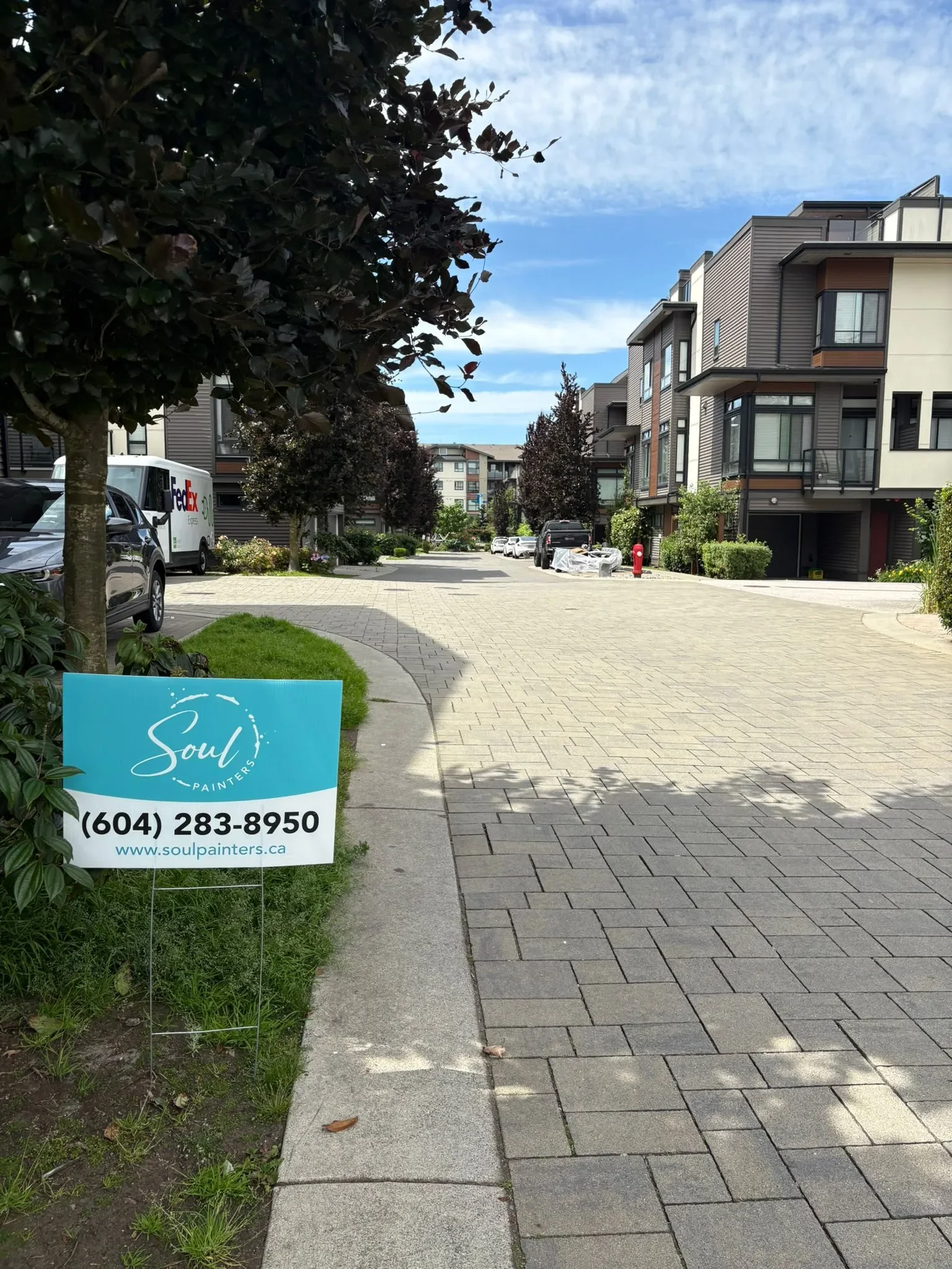 A residential street with modern apartment buildings, parked cars, a sidewalk, trees, and a sign for Soul Painters with contact information.