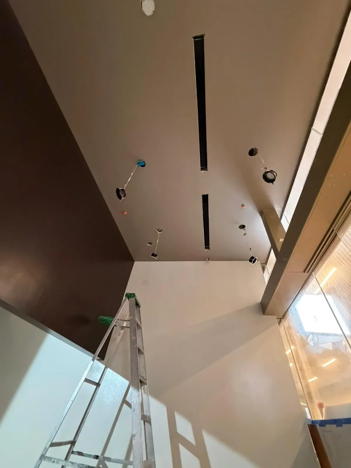 Ceiling under construction with lighting fixtures, ladder, and window glare.