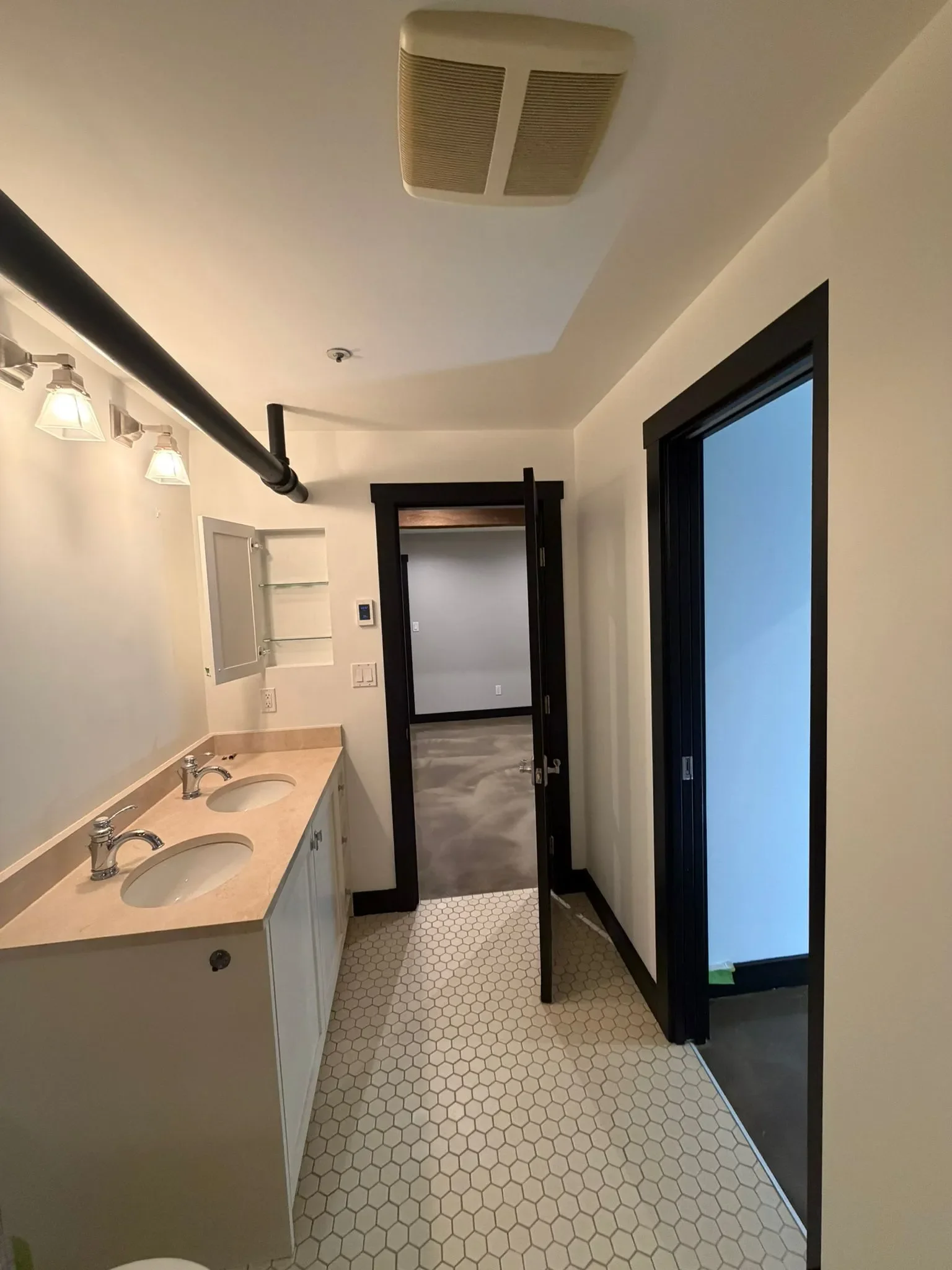 A bathroom with a double sink vanity, a black framed door, a frosted glass shower, hexagon white tile flooring, and a white ceiling with a vent and a black pipe in Richmond.
