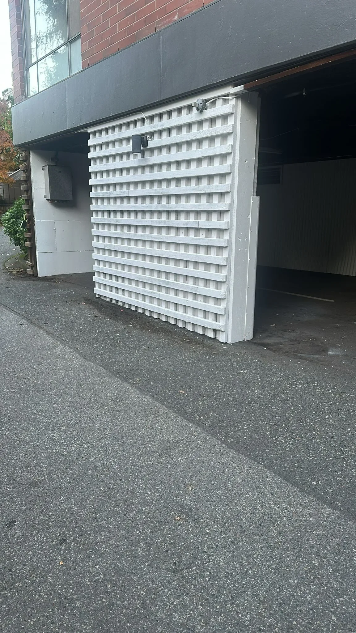 Parking garage entrance with a white lattice facade, gray asphalt driveway, and a brick and painted wall building