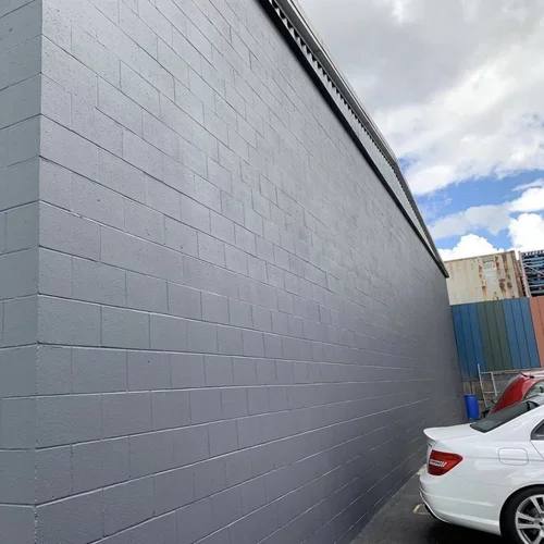 Large gray concrete block wall with car parked next to it in a parking lot under blue sky with clouds, painted and maintained by painters offering exterior painting services and free estimate near you.