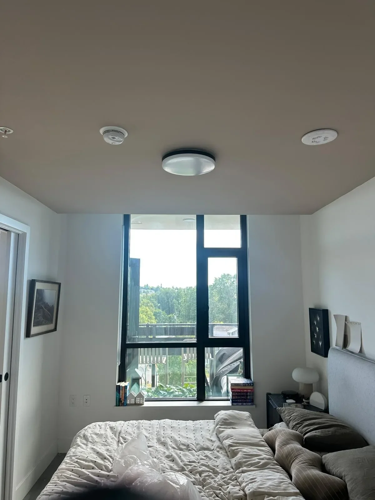 A bedroom with a large window showing a view of trees and a balcony outside. There is a bed with white bedding, pillows, and a brown blanket. A small black nightstand with a lamp and stacked books is beside the bed. Two framed pictures are on the wal