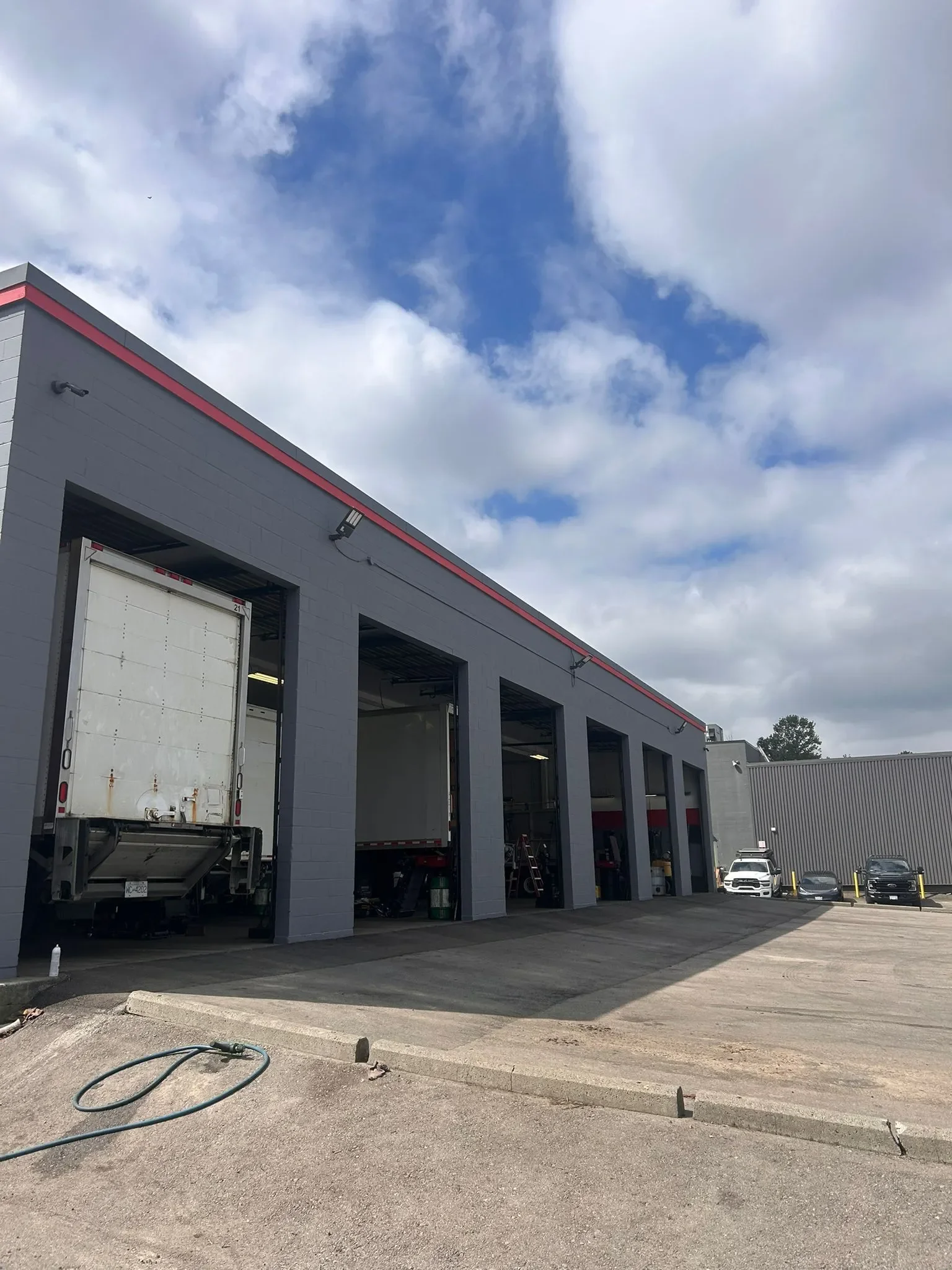 A commercial building with multiple garage bays, some of which are open, and several parked cars outside. The sky is partly cloudy with patches of blue.