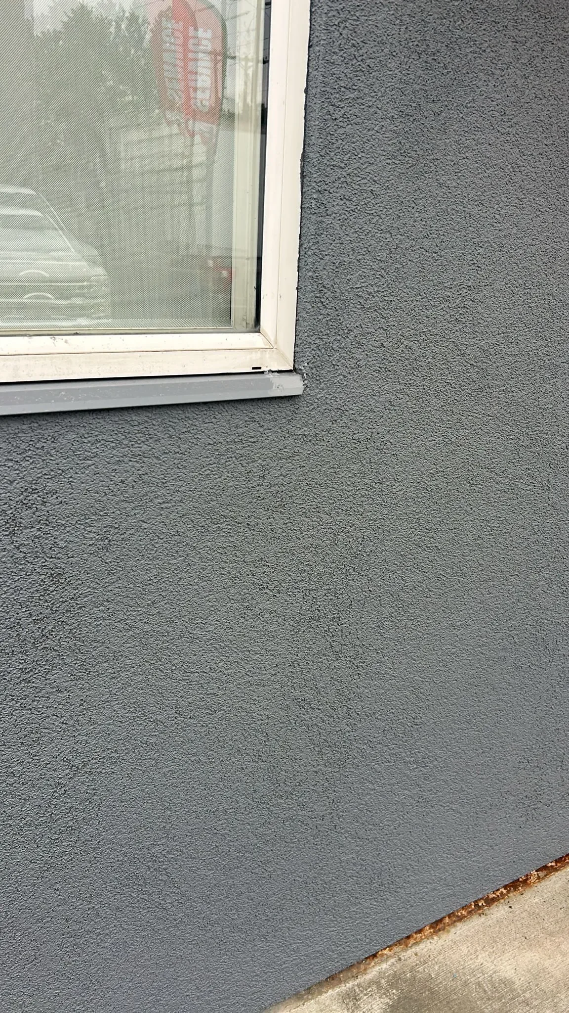 Close-up view of a building's exterior wall with a small window, showing the textured gray surface of the wall and part of the window frame.
