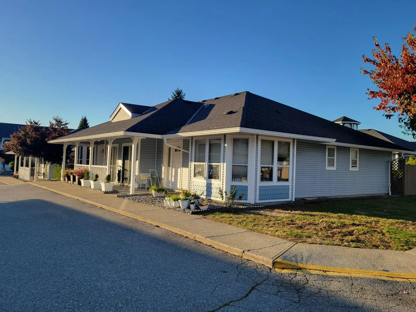 Single-story house with a porch decorated with potted plants, white trim, and gray exterior, illuminated by late afternoon sunlight, in a residential neighborhood.