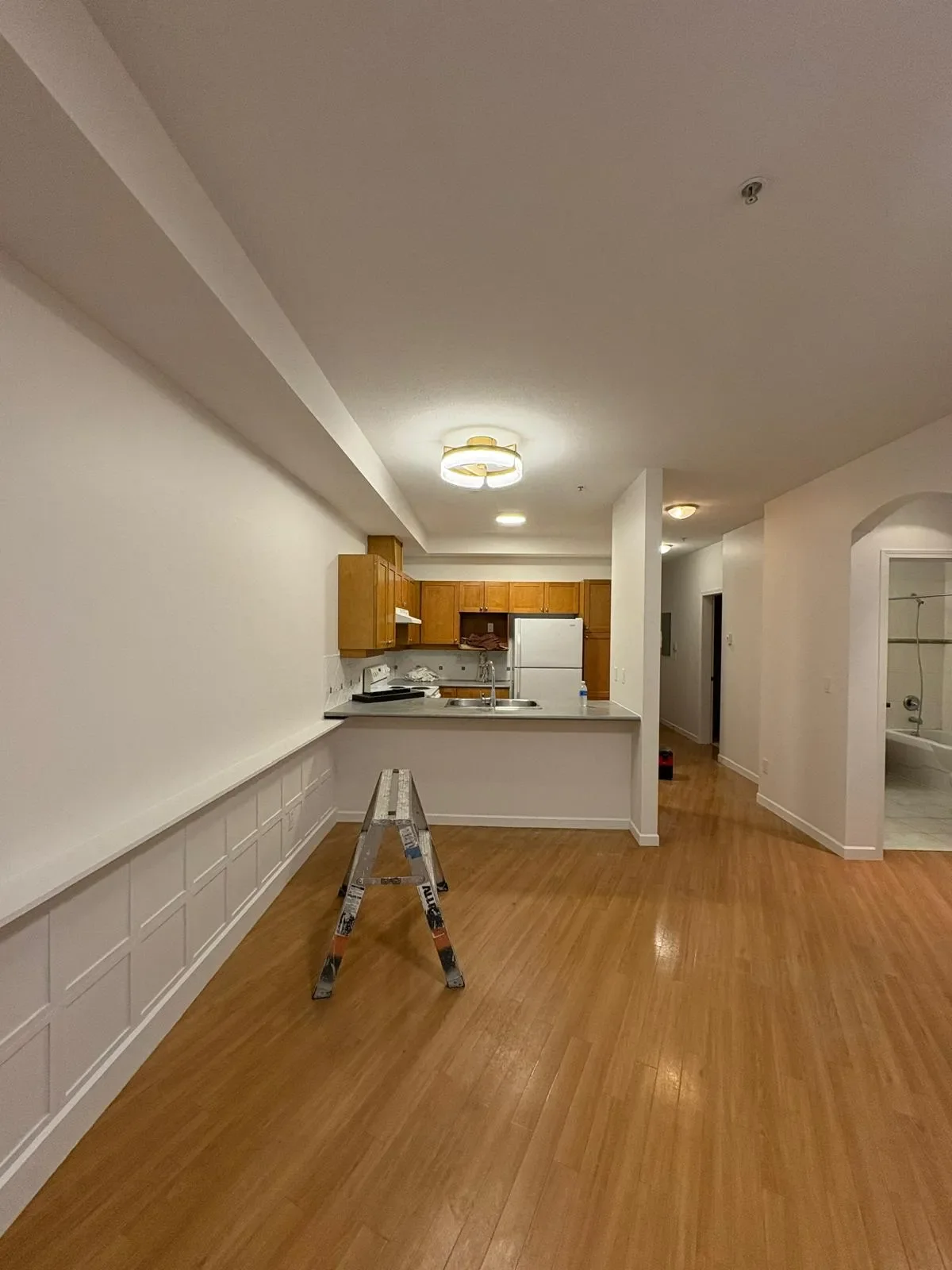 Empty apartment kitchen with wooden cabinets, white appliances, a small ladder in front, and hardwood floors.