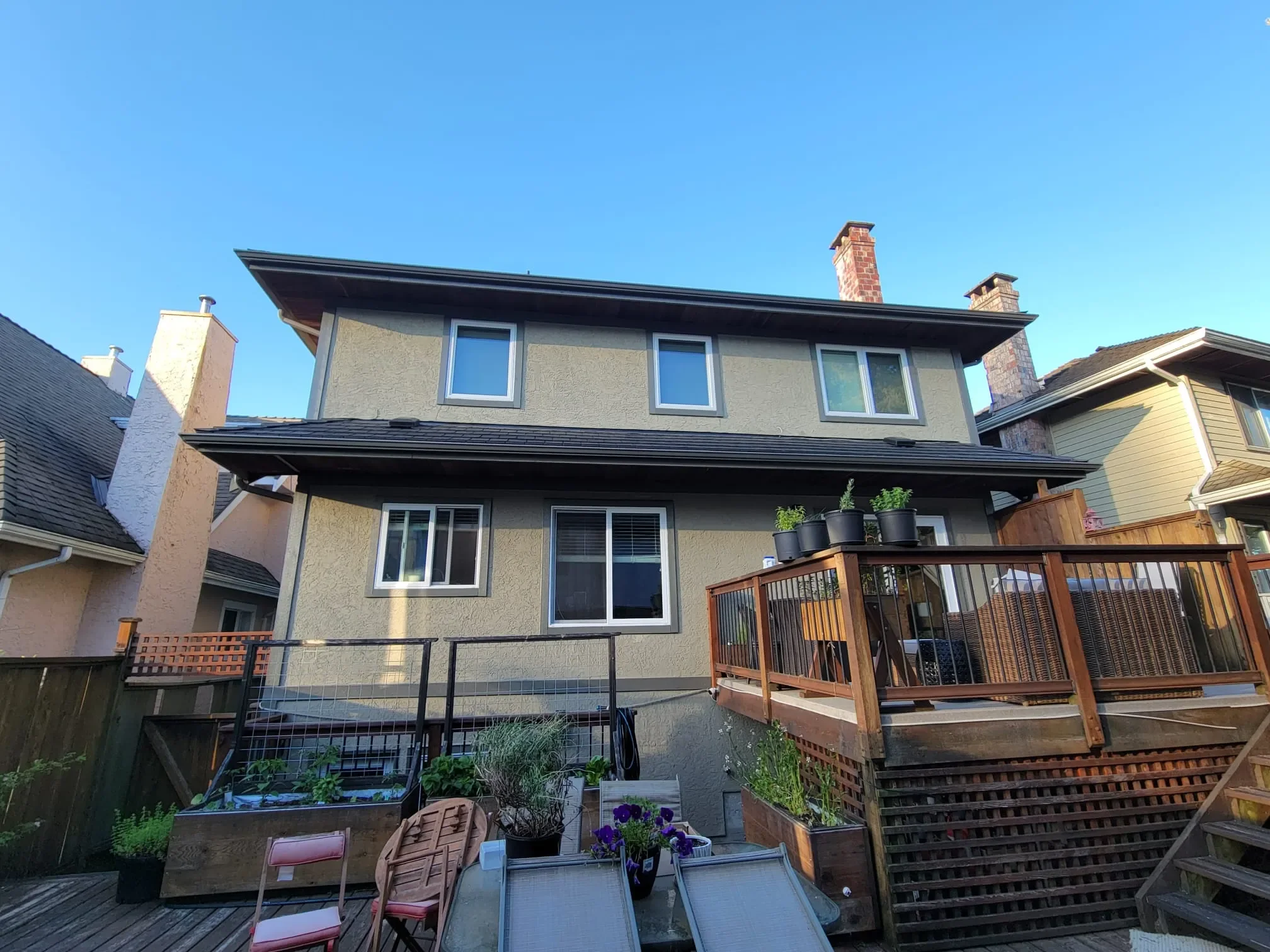 View of a backyard with a multi story house, wooden deck, garden beds, potted plants, and outdoor furniture under a clear blue sky in Maple Ridge.