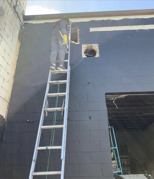Person on ladder painting exterior wall of building dark blue, wearing gray pants, dark hoodie, yellow gloves, and safety harness, with small window or vent near top, painted and maintained by painters offering exterior painting services and free est