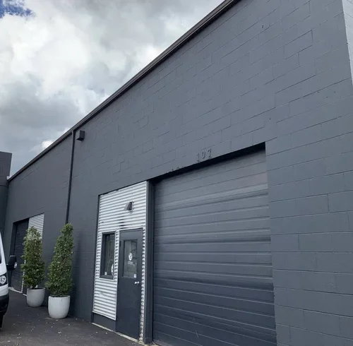 Painters working on the exterior of a gray industrial building with large rolling door, small door, and potted bushes, offering free painting estimate and exterior painting services near you.