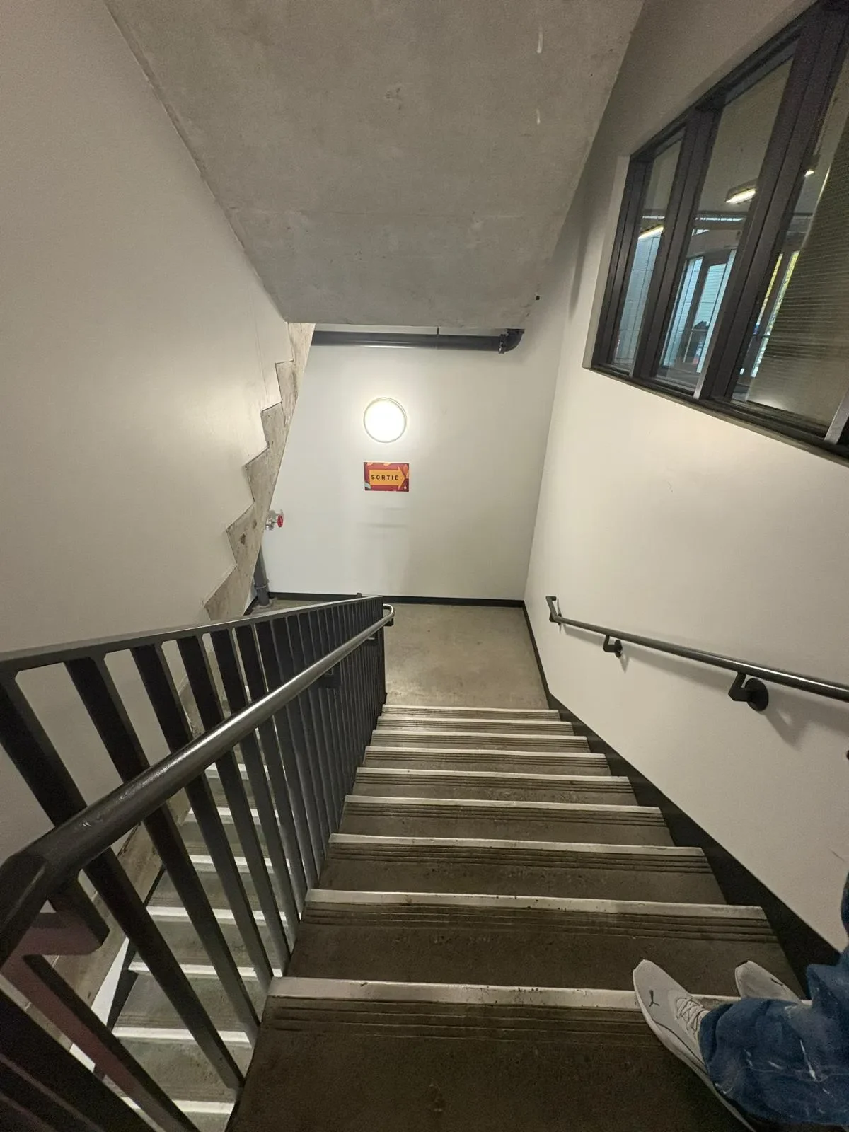 Interior view of a stairway descending to a lower floor, with black handrails on both sides, and a 'Sortie' exit sign on the wall at the bottom. Part of a person's white shoe and blue jeans are visible in the lower right corner.
