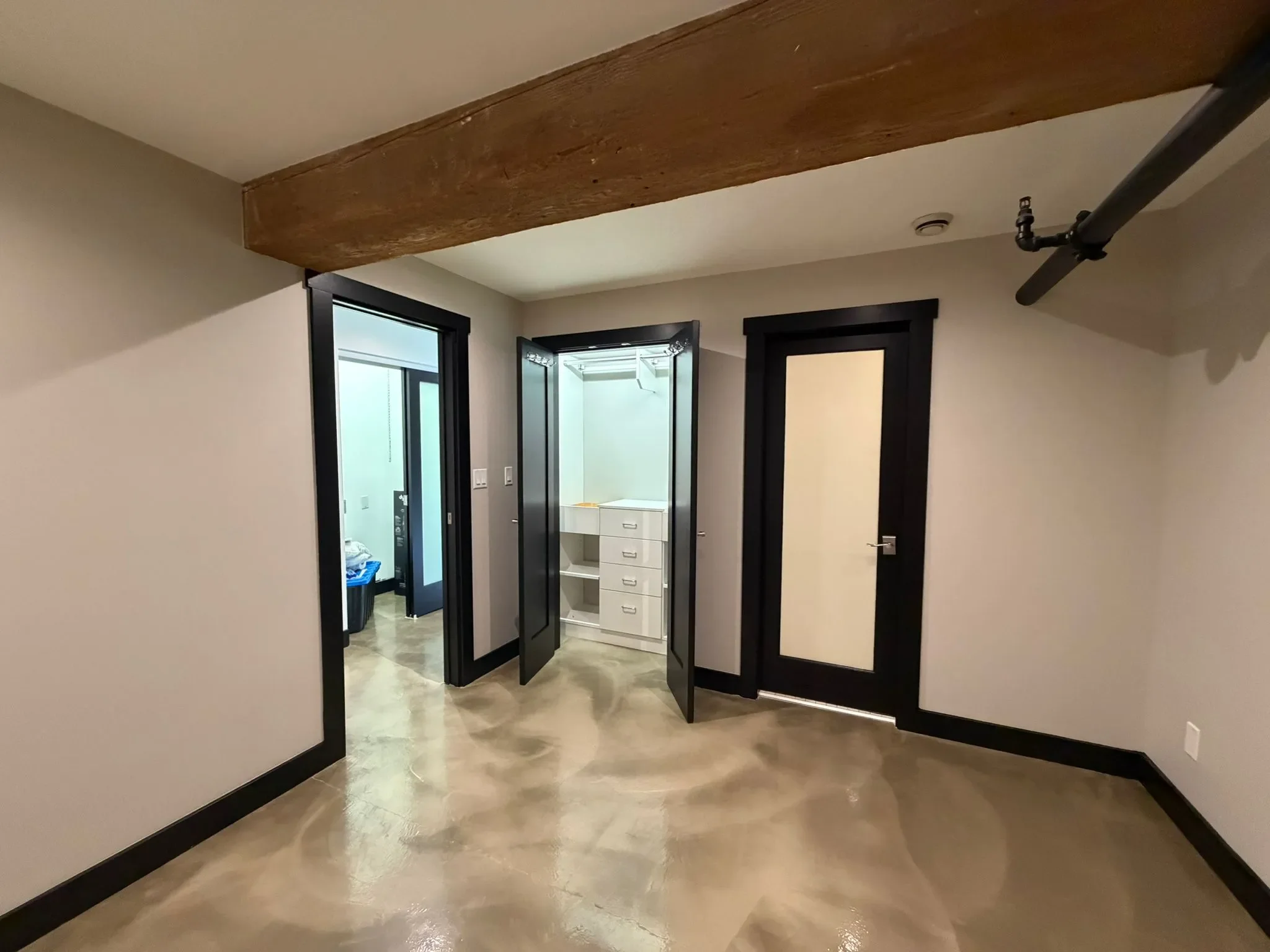 Interior of a modern building with concrete flooring, black doorframes, and a ceiling beam in Richmond. Open closet with white drawers and a mirror, adjacent to a black door, leads to another room with a glass door closet and laundry items. Pipes are