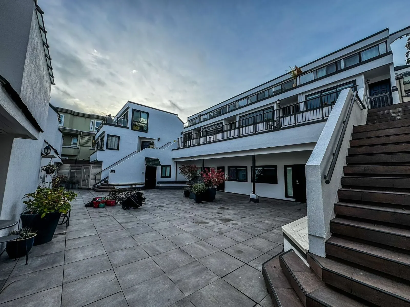 Apartment complex courtyard with stairs, potted plants, and outdoor furniture under a cloudy sky.