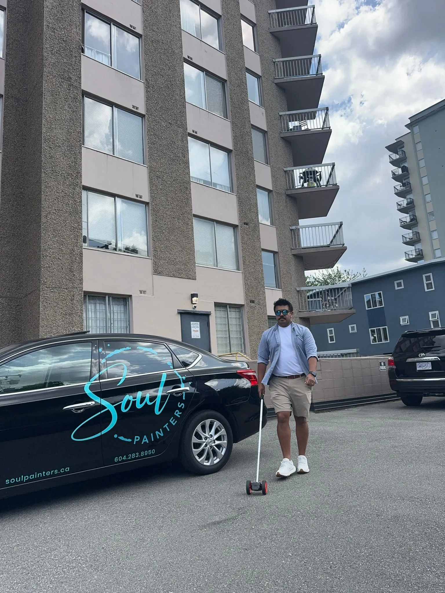 A man stands on a parking lot in front of a black car with the logo 'Soul Painters'. He is wearing sunglasses, a light blue shirt over a white t-shirt, beige shorts, and white sneakers. The background features multi-story apartment buildings with bal
