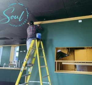 Person on yellow ladder painting interior wall in a room, with neon "Soul Paint" sign on ceiling, painted and maintained by painters offering interior painting services and free estimate near you.