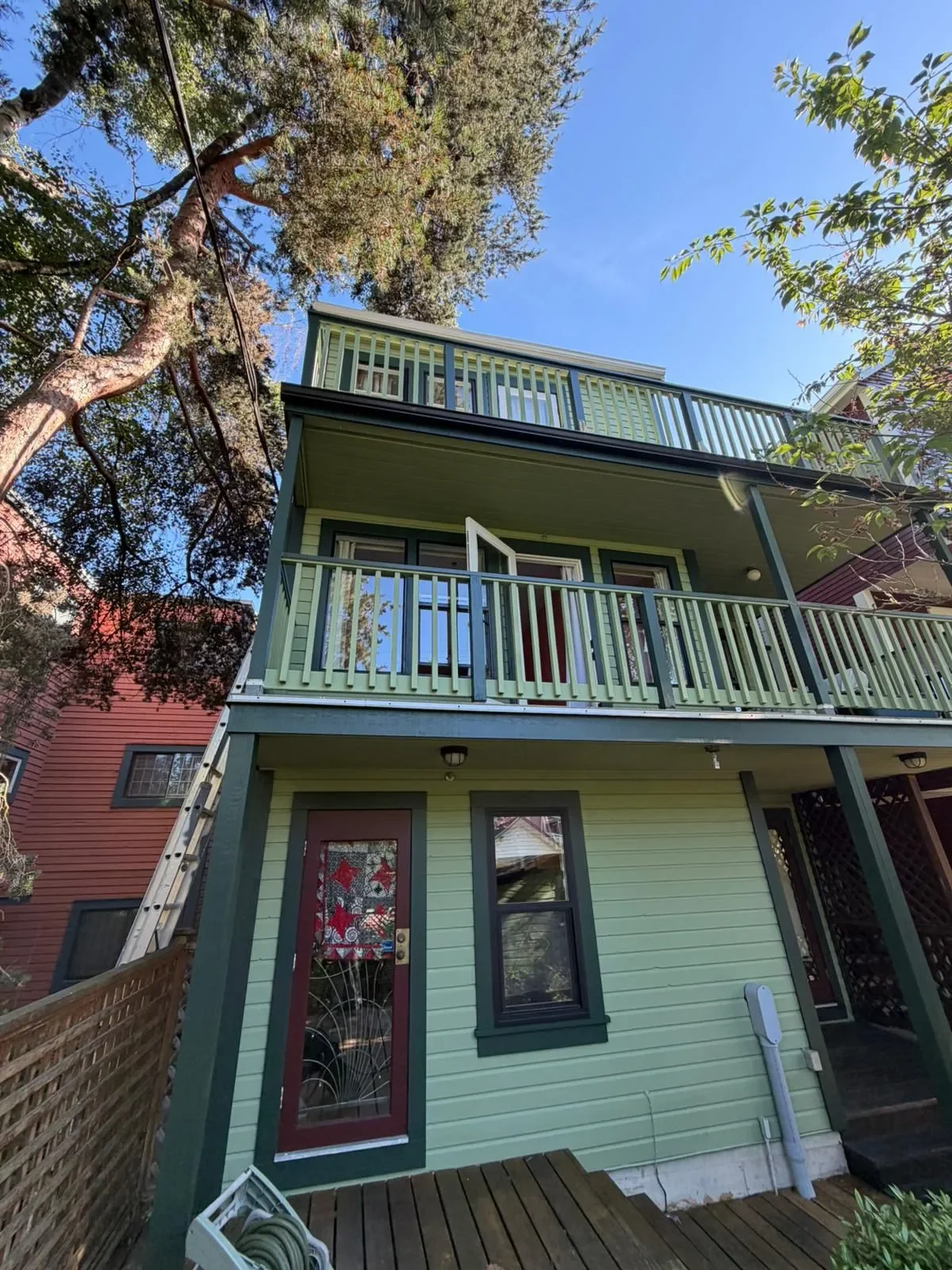 A three story house with green and black exterior, featuring a small balcony on the second and third floors, a decorative door, a ladder leaning against the house, and trees surrounding it under a clear blue sky in Surrey.