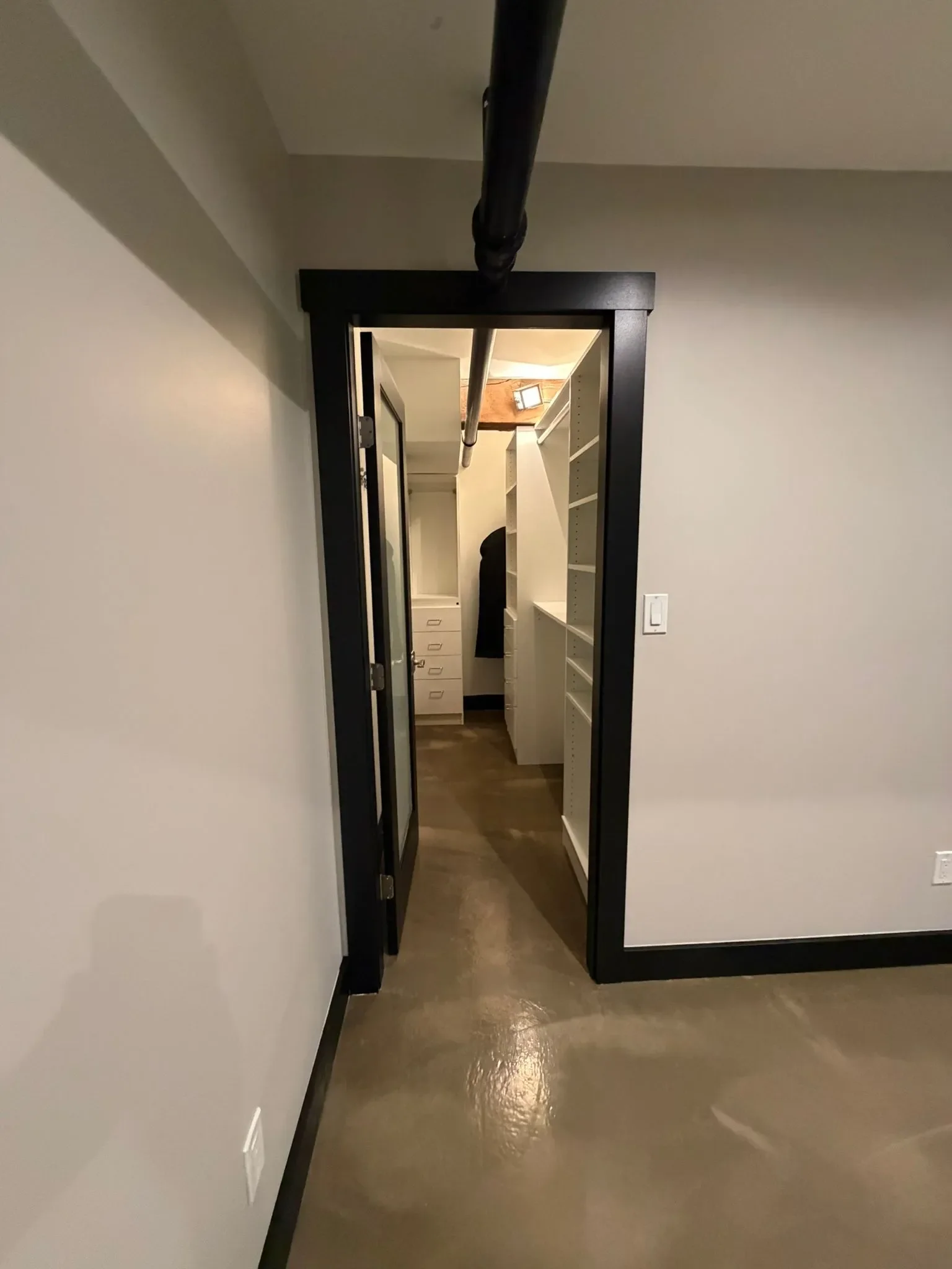 A narrow hallway with concrete floor, white walls, and a black doorframe leading to a walk-in closet with white shelves and drawers in Richmond.