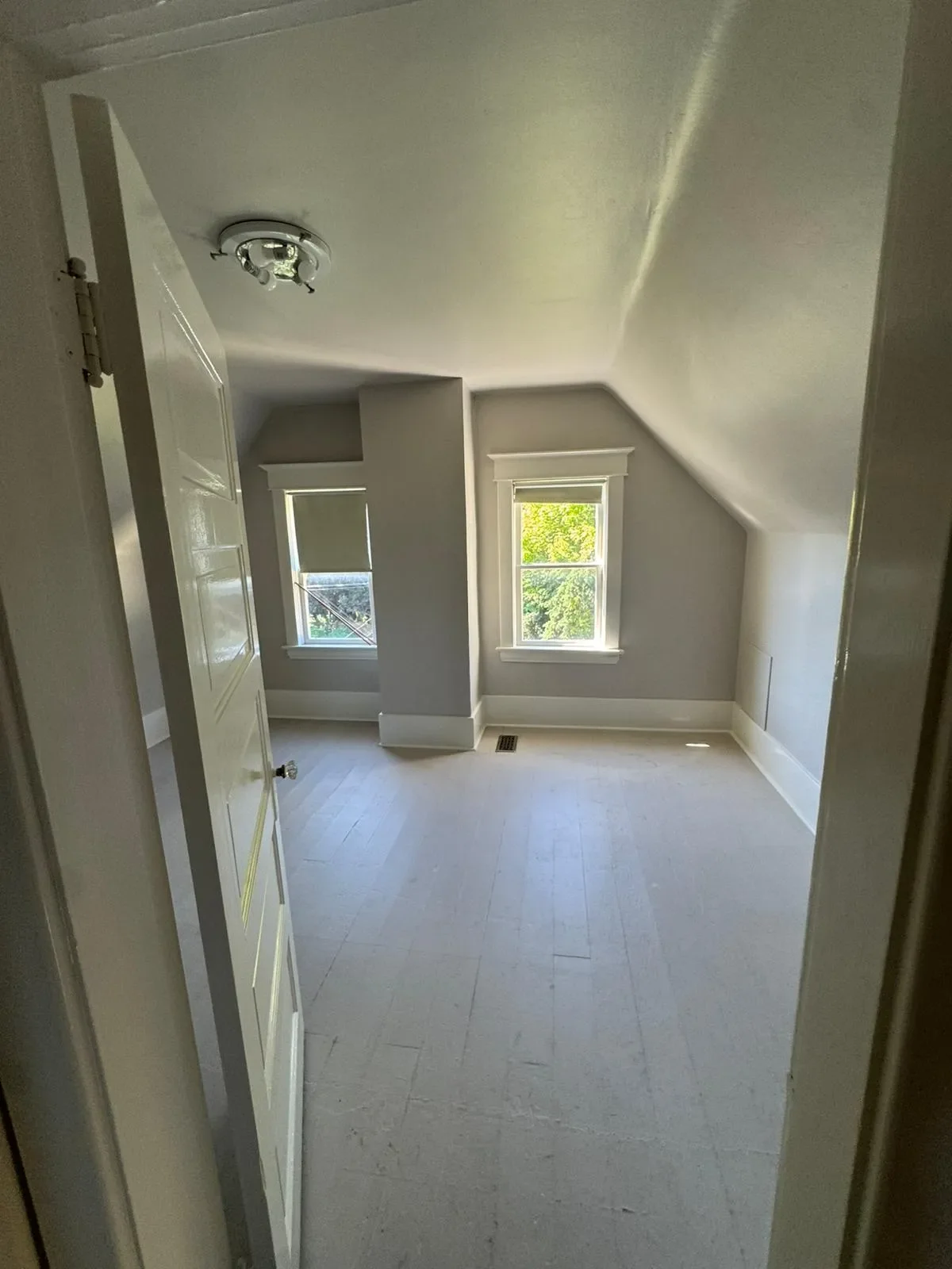 An empty room with three windows, light colored walls, white trim, and a sloped ceiling in Langley. There is a door partially open on the left side. The room has a floor vent near the windows and natural light coming through the windows.