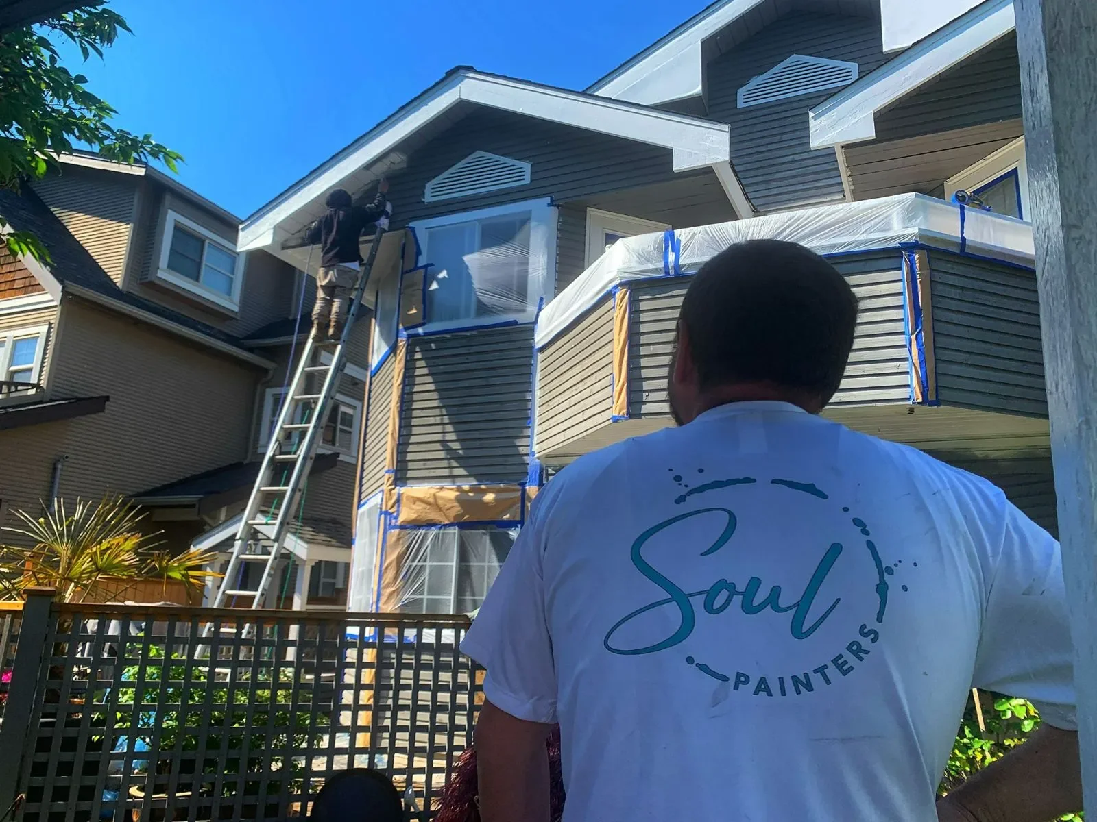People painting the exterior of a house, one person on a ladder painting near the roof, another person watching from below. The house has beige siding and a balcony with a white railing, and the sky is blue.