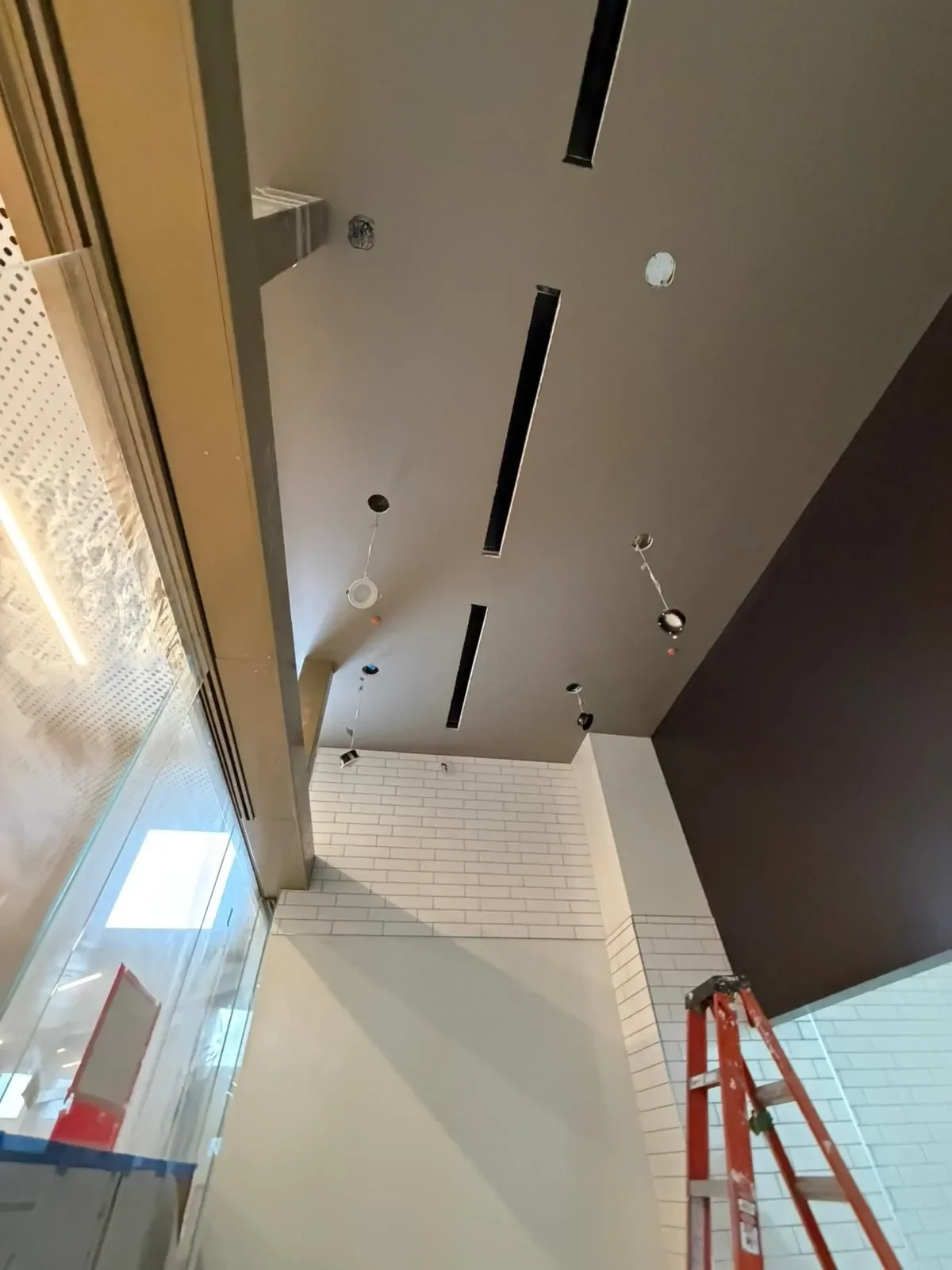 Ceiling of a building under construction or renovation, with black linear vents and hanging electrical wires, adjacent to white brick walls and a glass wall with a frame.