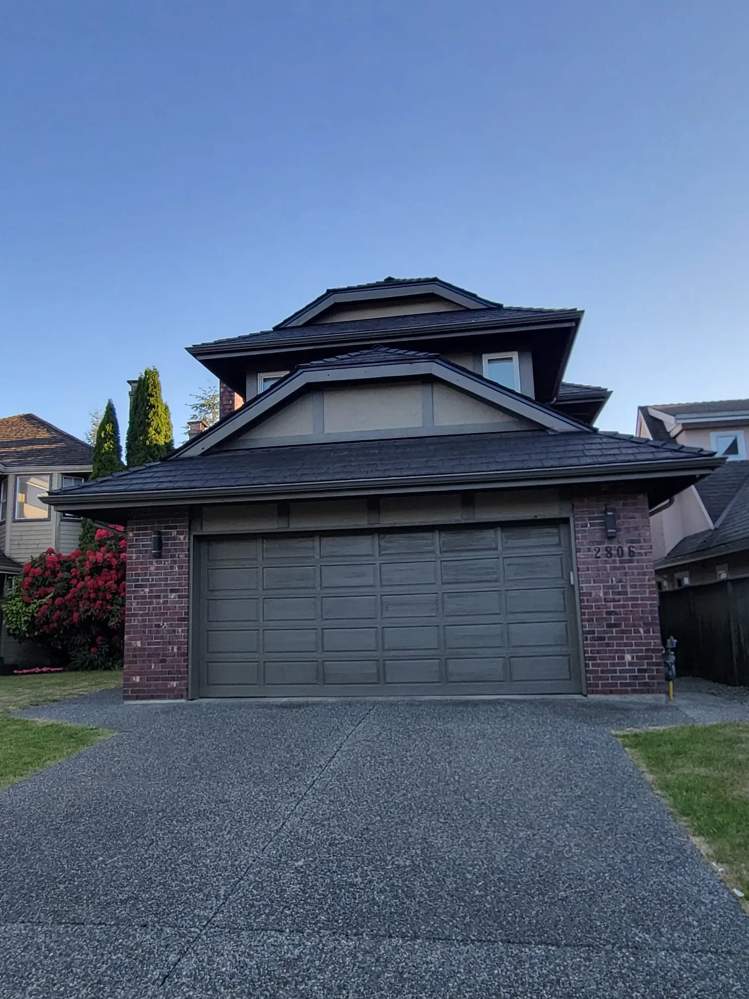 A two-story house with a brick exterior and a gray garage door, surrounded by a driveway, green lawn, and some flowering bushes, under a clear blue sky in Belcarra.