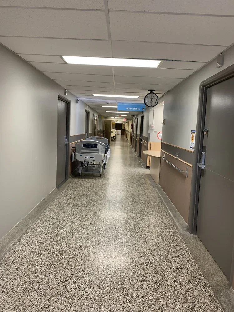 Long hospital corridor with patient beds, medical equipment on the left, nurse station sign, wall clock showing 8:11, beige and gray walls, and speckled gray floor, painted and maintained by painters offering free interior painting estimate near you.