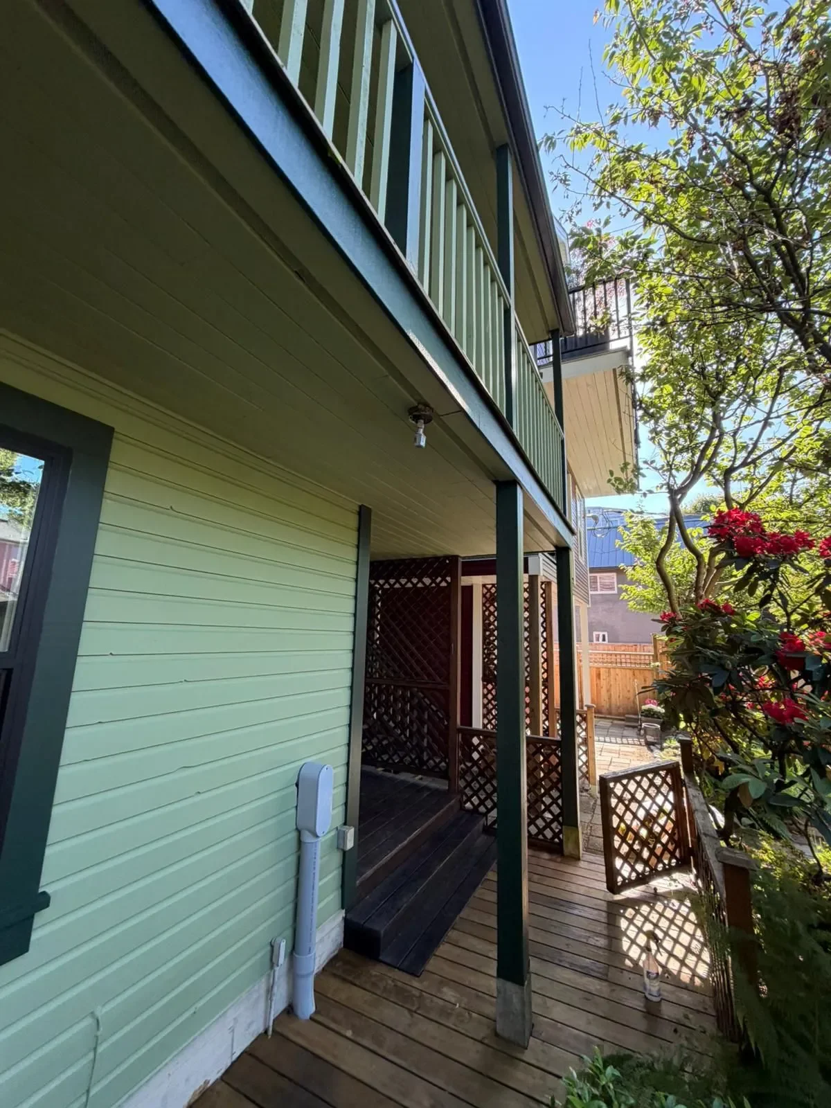 A two-story house with green siding, second-floor balcony with green railing, and an outdoor wooden deck area with a lattice privacy screen, surrounded by trees and flowering plants under a clear blue sky in Surrey.