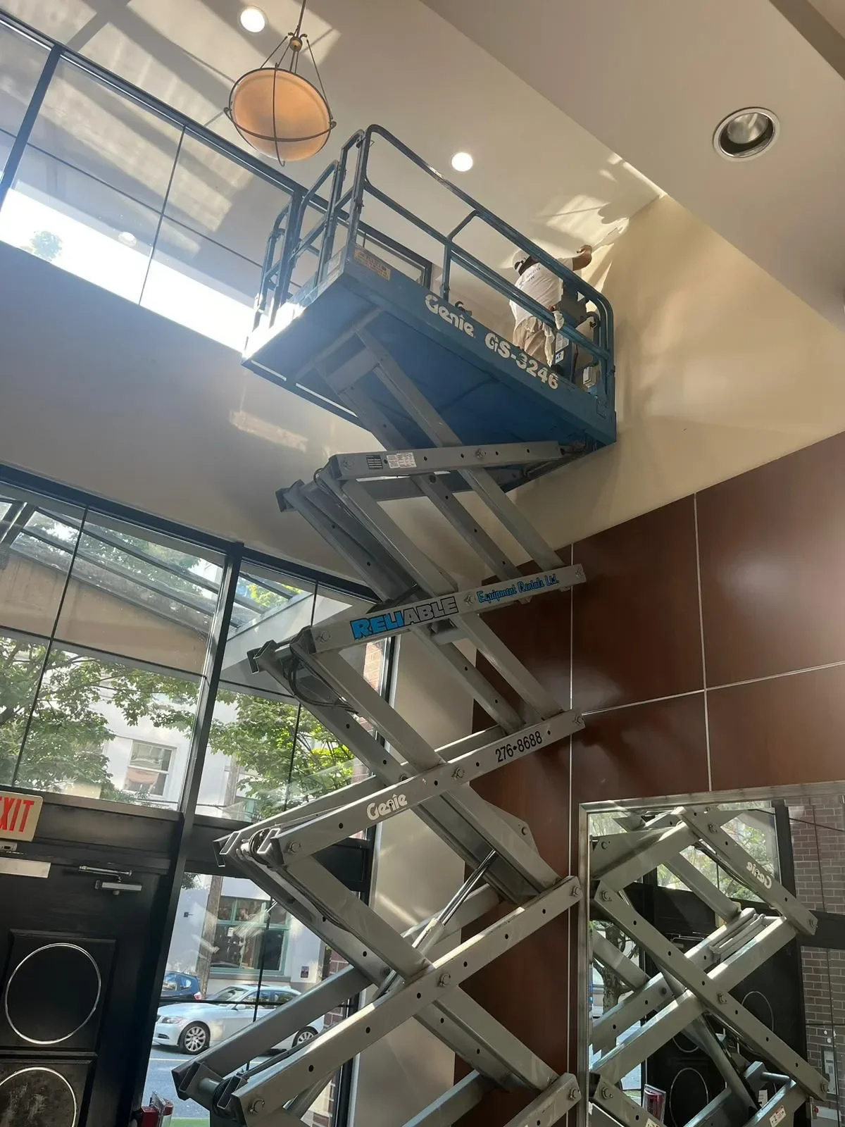 A worker on a blue Genie lift working on the ceiling of a modern building's interior, with large windows and natural light.