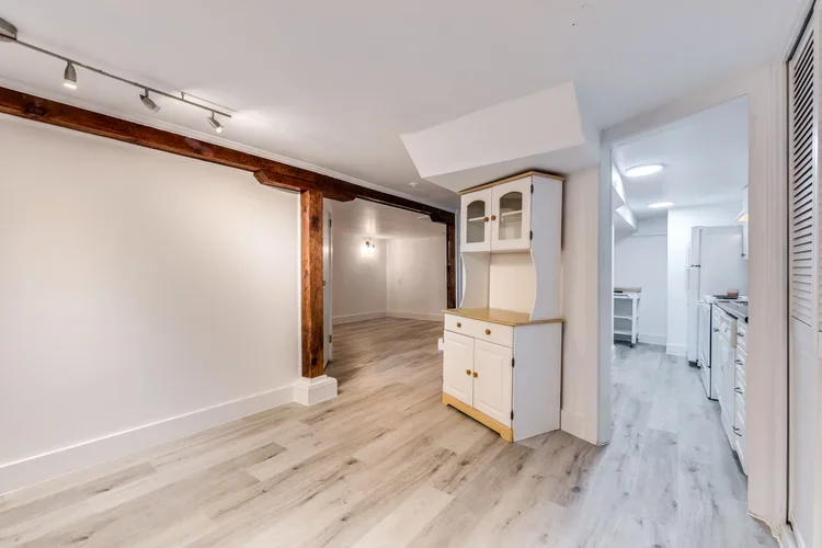 Empty living space with wood beams, white walls, light wood flooring, and a white cabinet near the kitchen opening to the kitchen with white appliances and cabinetry.