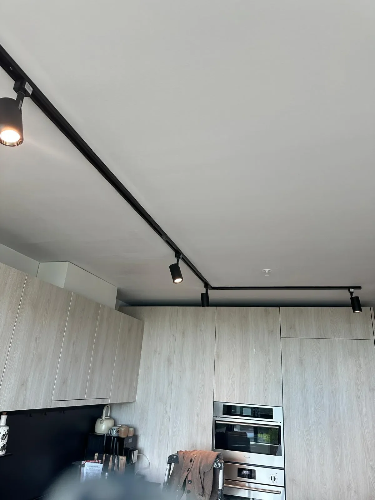 Modern kitchen ceiling with black track lighting and spotlights, wood cabinets, and built-in oven in Coquitlam.