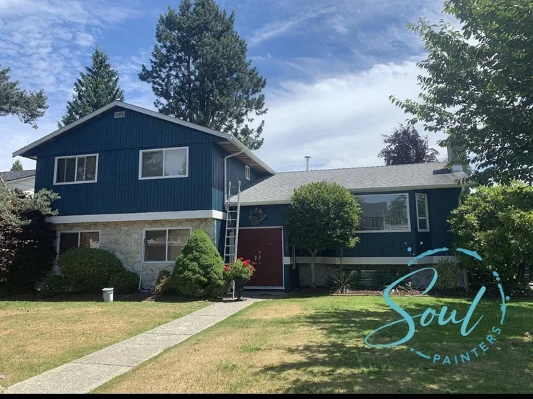 A two-story house with a blue exterior, stone foundation, and a gray roof, surrounded by trees and a lawn, under a partly cloudy sky.