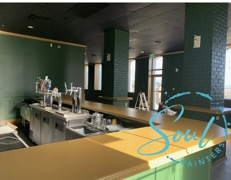 Interior of bar or restaurant under construction with dark green walls, large windows, long yellow countertop, bar area with beer taps and glasses, and "Soul Painters" logo overlaid, painted and maintained by painters offering free commercial paintin
