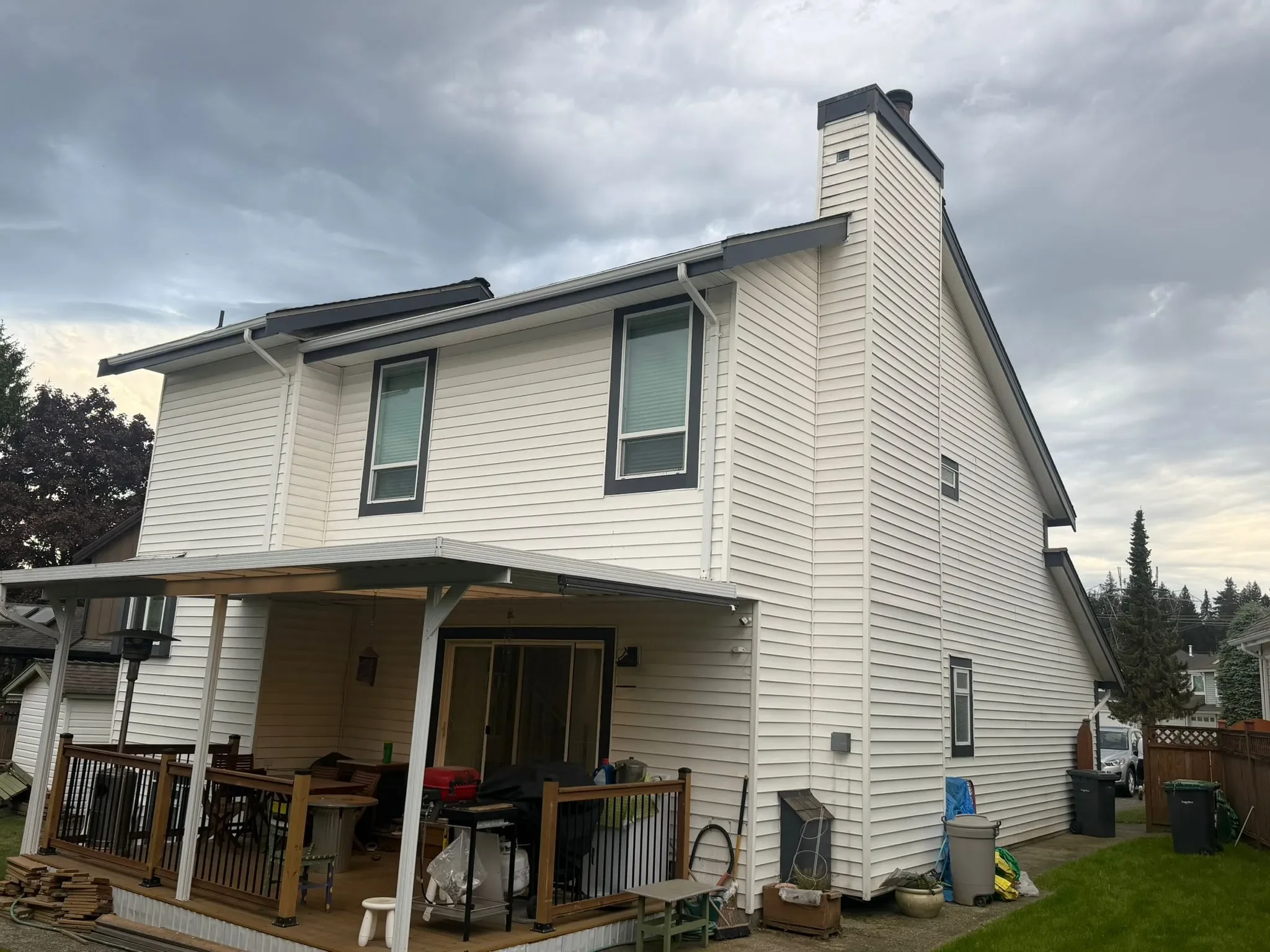 The photo shows the exterior of a two-story white house with a wood deck and patio furniture in the backyard in North Vancouver. The house has several windows, a chimney, and a sloped roof. The sky is cloudy, and there are trees and other houses in t